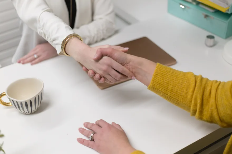 Dos personas se dan la mano sobre una mesa de despacho blanca con una tablet y una taza de cafe